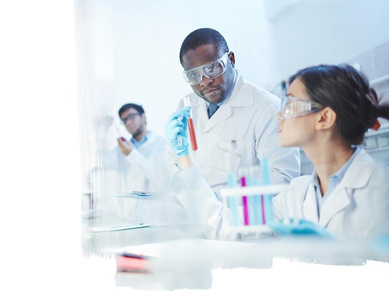Female Asian laboratory scientist in lab coat and safety goggles showing test tube with red liquid to curious African-American colleague in laboratory. Latin-American scientist in background.